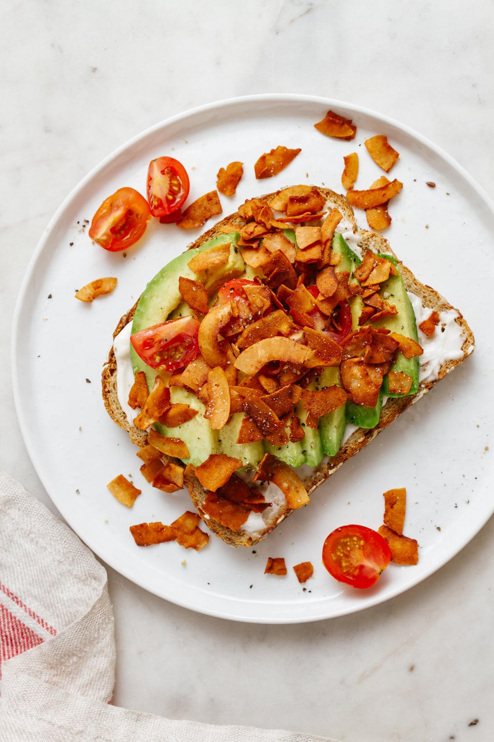 top down view of toast with sliced avocado, coconut bacon, tomato, salt and pepper on a small white plate.