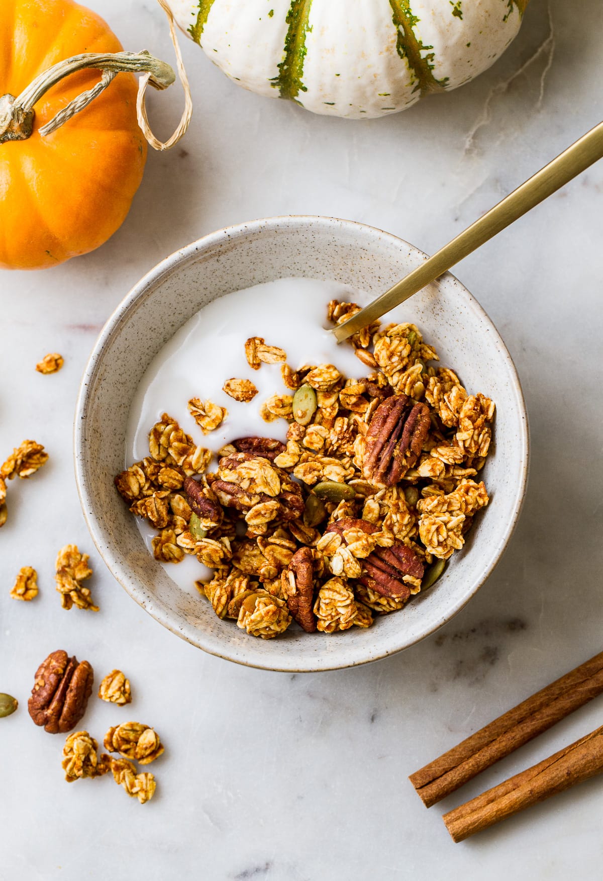 top down view of a bowl with a serving of coconut yogurt and pumpkin granola with a gold spoon surrounded by items.