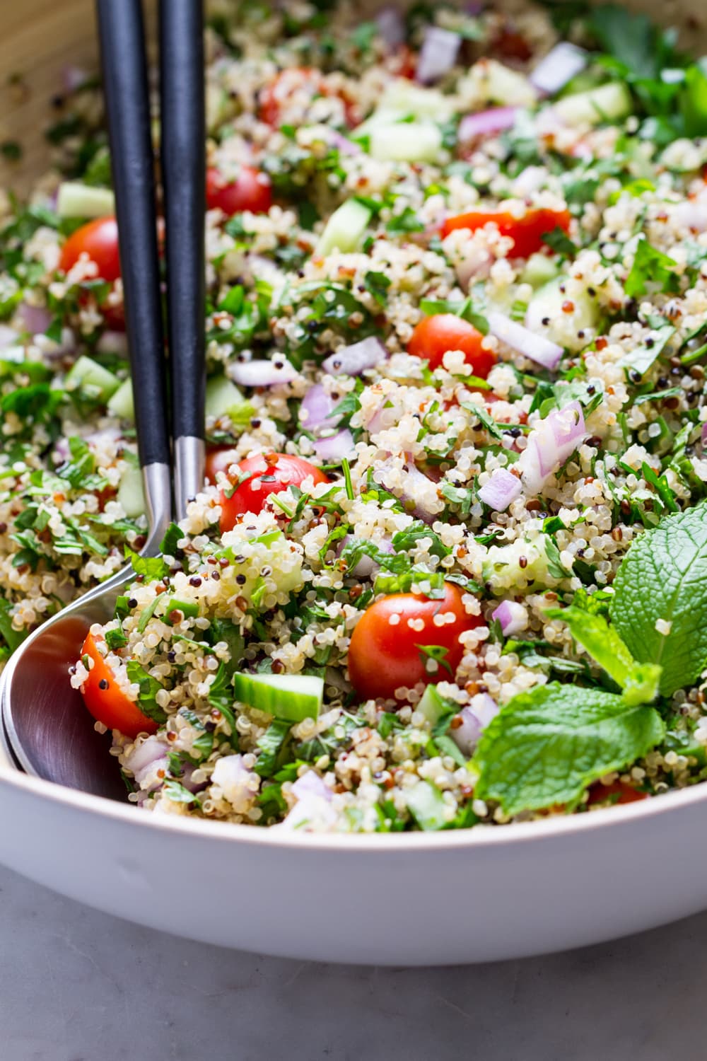 up close, side angle view of healthy quinoa tabbouleh in a large mixing bowl with serving utensils.