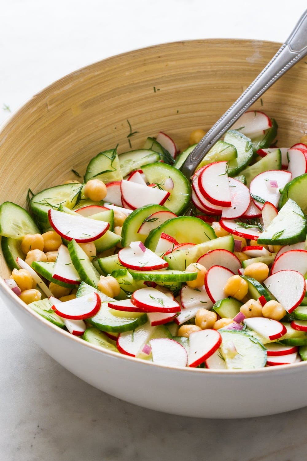 side angle top down view of a white bowl serving bowl filled with radish and cucumber salad with a serving spoon on a marble slab
