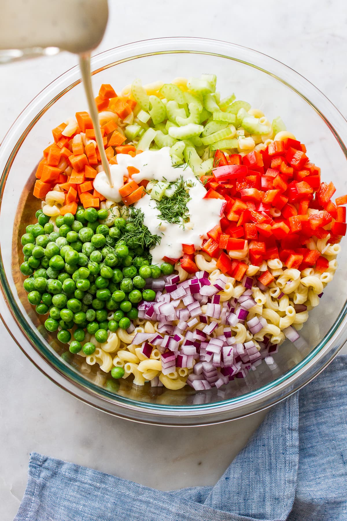 top down view of ingredients to make vegan macaroni salad in a glass bowl with creamy dressing being poured overtop.
